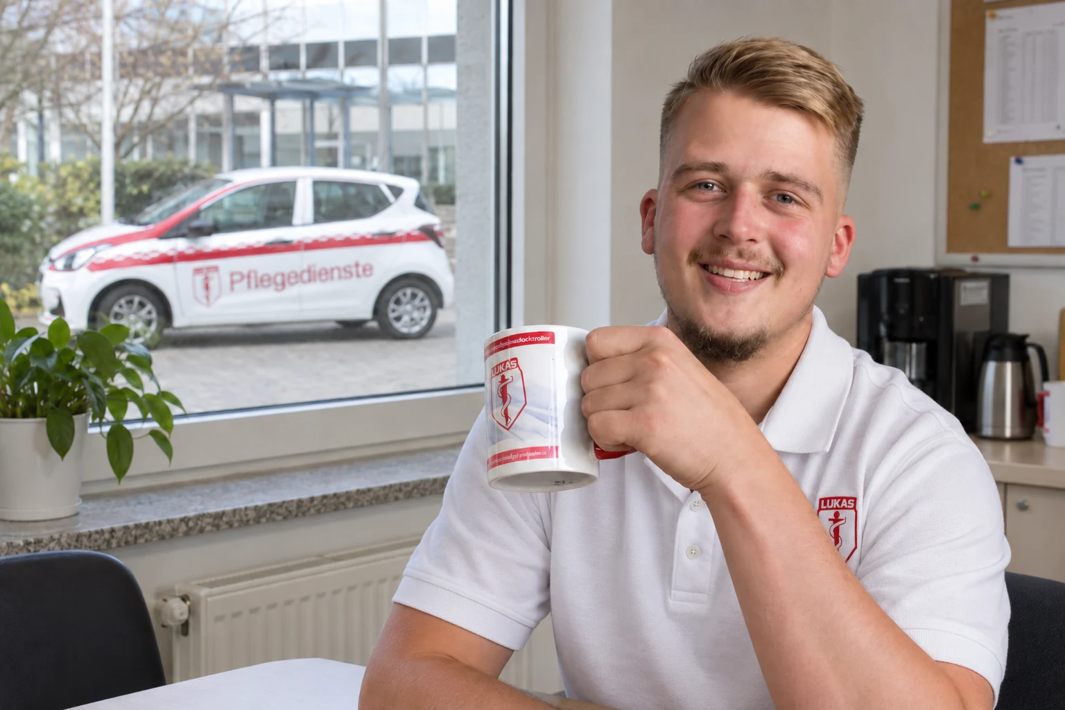 Ein Mitarbeiter von St. Lukas sitzt mit einer Tasse am Fenster; im Hintergrund steht ein Pflegefahrzeug.